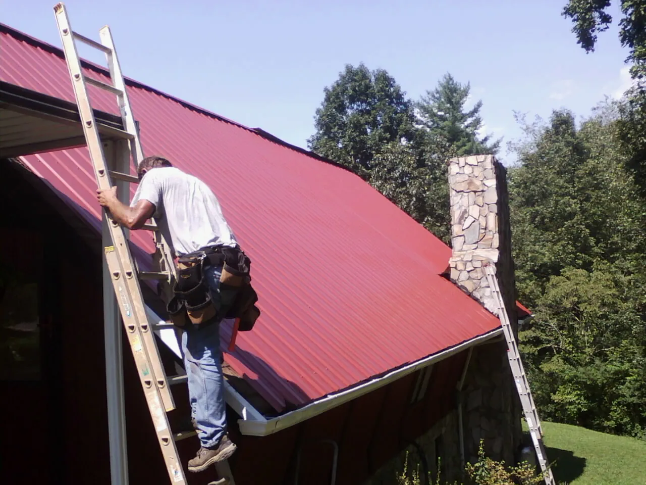 Roofer on a ladder beside a red metal roof and stone chimney — Soffit & Fascia Repair by Roofing Brothers in Battle Creek